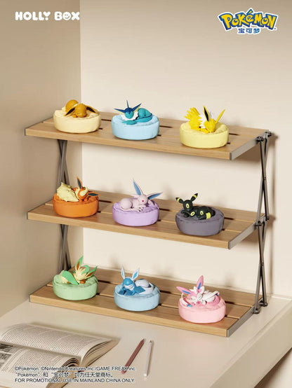 Nine Pokémon Eeveelution figures sleeping on colorful, soft-textured beanbag cushions displayed on a wooden shelf.
